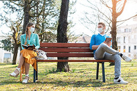 Junge Frau und junger Mann sitzen auf einer Parkbank mit Abstand und Maske