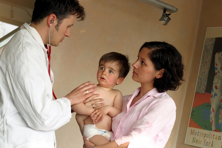 Eine Frau hält ein Kleinkind auf dem Arm, während es von einem Arzt untersucht wird.