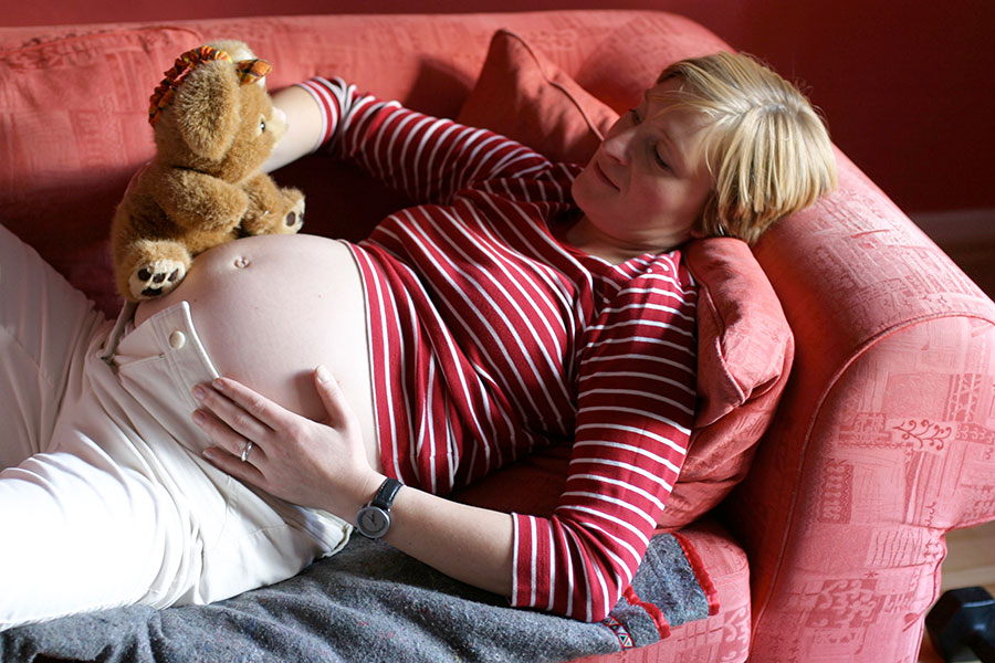Hochschwangere junge Frau liegt auf Sofa und hält Teddy in der Hand.