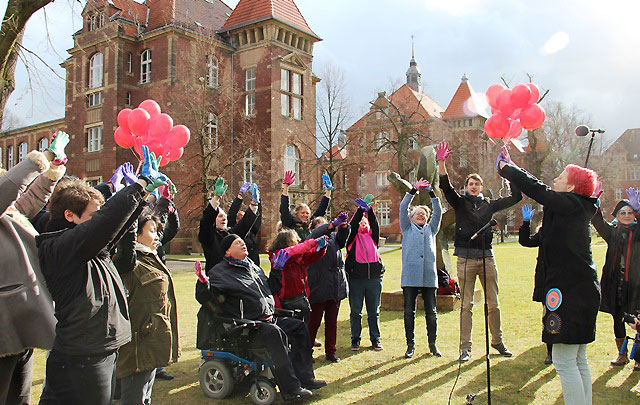 Eine Gruppe von Menschen, die bunte Handschuhe tragen, lassen rote Luftballons steigen. 