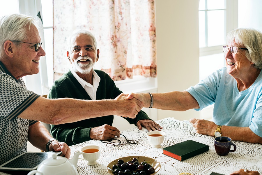 Drei ältere Menschen sitzen zusammen an einem Tisch, zwei davon schütteln sich die Hände.