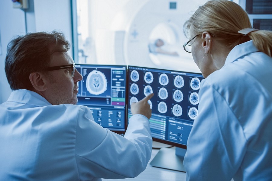 Ein Arzt sitzt vor Bildschirmen, auf denen Aufnahmen eines Gehirns zu sehen sind und und zeigt etwas einer Ärztin, die rechts neben ihm steht