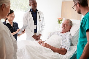 Ein älterer mann liegt in einem Krankenbett, mehrere Menschen in verschiedenfarbiger Krankenhauskleidung stehen um ihn herum