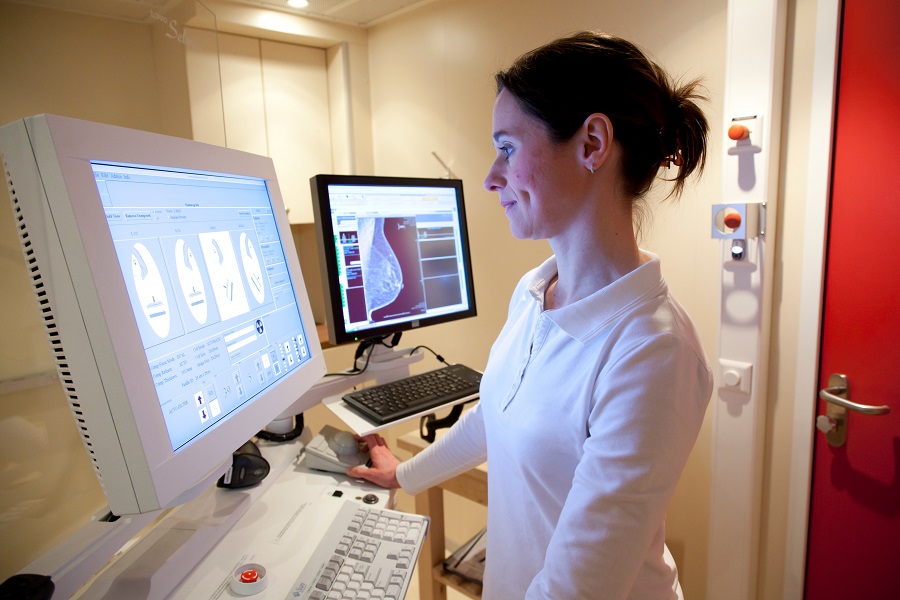 Eine Frau steht vor einem Computerbildschirm, auf dem Daten abgebildet sind, rechts neben ihr steht ein Bildschirm mit einer Aufnahme einer weiblichen Brust