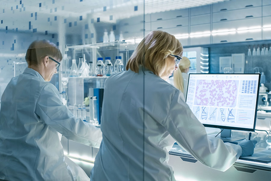 Zwei Forscherinnen in weißem Kittel sitzen an der Laborbank vor verschiedenen Proben in Glasröhrchen und Reagenzgläsern. Vor der rechten Forscherin steht ein Computer, auf dessen Bildschirm Mikroskopaufnahmen biologischer Proben zu sehen sind. 