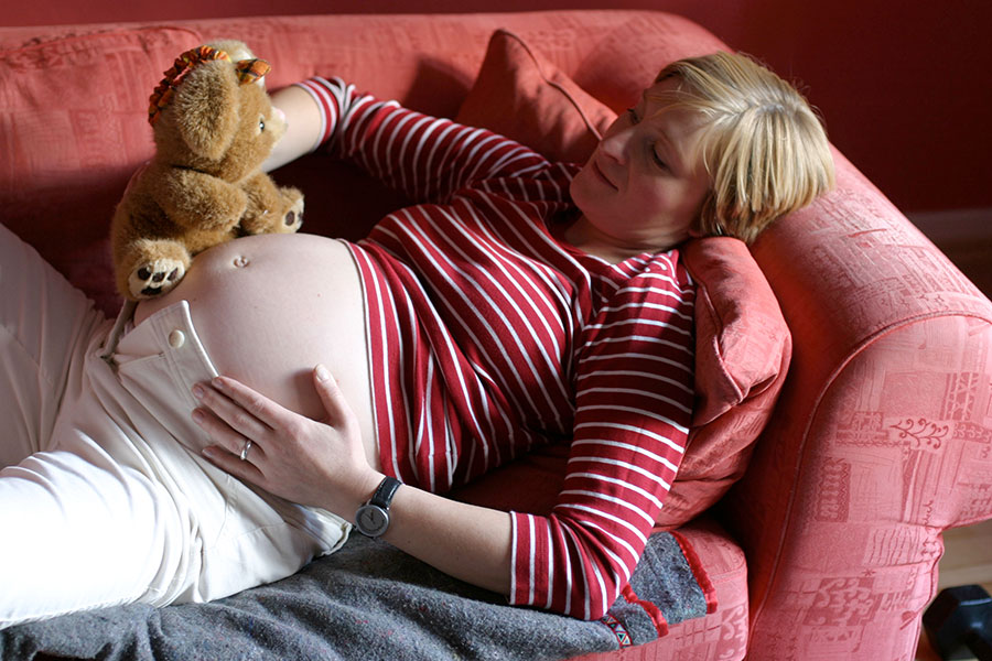Eine schwangere Frau liegt auf einem Sofa und hält ein Kuscheltier an ihren Bauch.