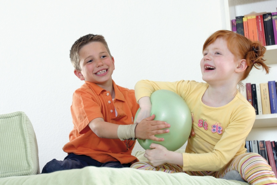 Eine Junge und ein Mädchen rangeln auf einem grünen Sofa lachend um einen grünen Ball. 