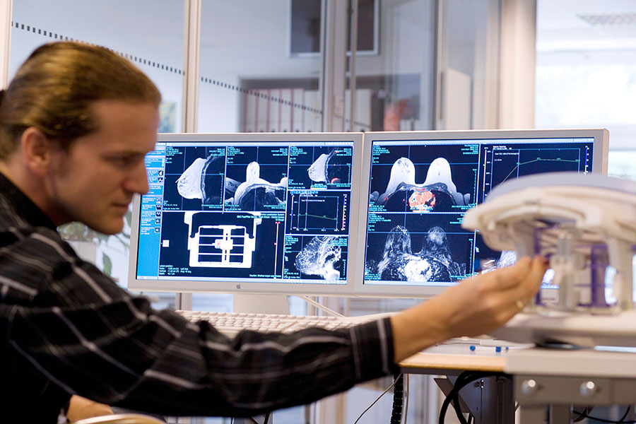 Wissenschaftler bedient ein Gerät mit Probengefäßen, im Hintergrund befinden sich Computermonitore auf denen MRT-Aufnahmen zu sehen sind.