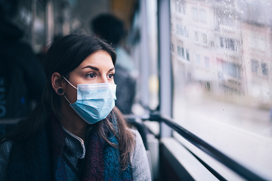 Junge Frau mit Mundschutz schaut aus dem Fenster eines fahrenden Busses auf verregnete Stadt