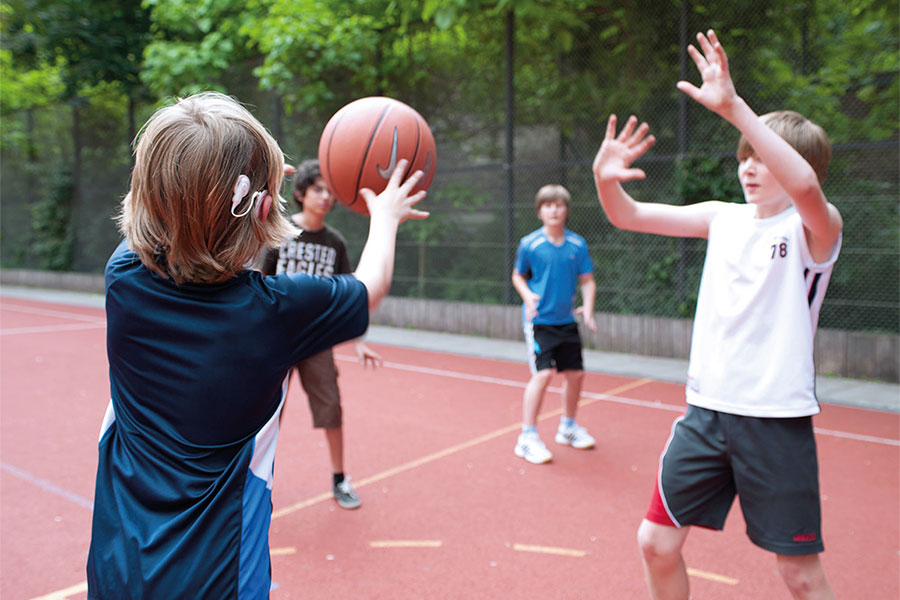 Kinder spielen mit Ball auf Sportplatz, wobei ein Junge ein Cochlea-Implantat hat und ein Hörgerät hinter dem rechten Ohr trägt.