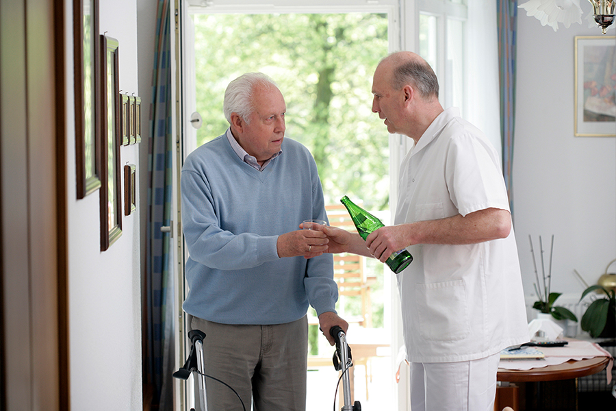 Pfleger reicht älterem Herrn in Pflegeeinrichtung ein Glas Wasser