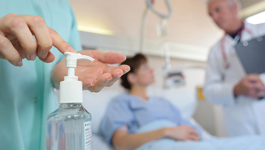 Medizinische Fachkraft betätigt Spender mit Desinfektionsmittel, während im Hintergrund Arzt mit einer Patientin spricht.