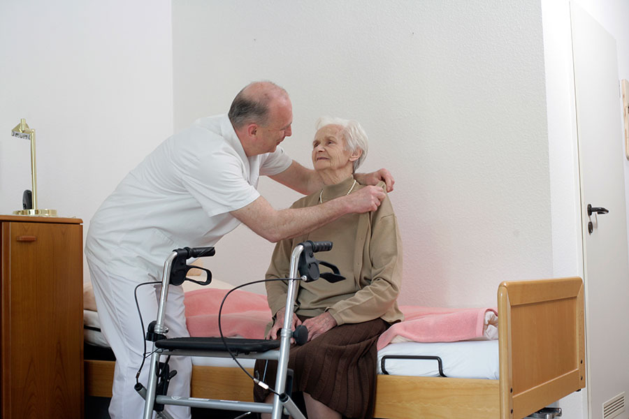 Ein Pfleger hilft einer älteren Dame eine Strickjacke anzuziehen.