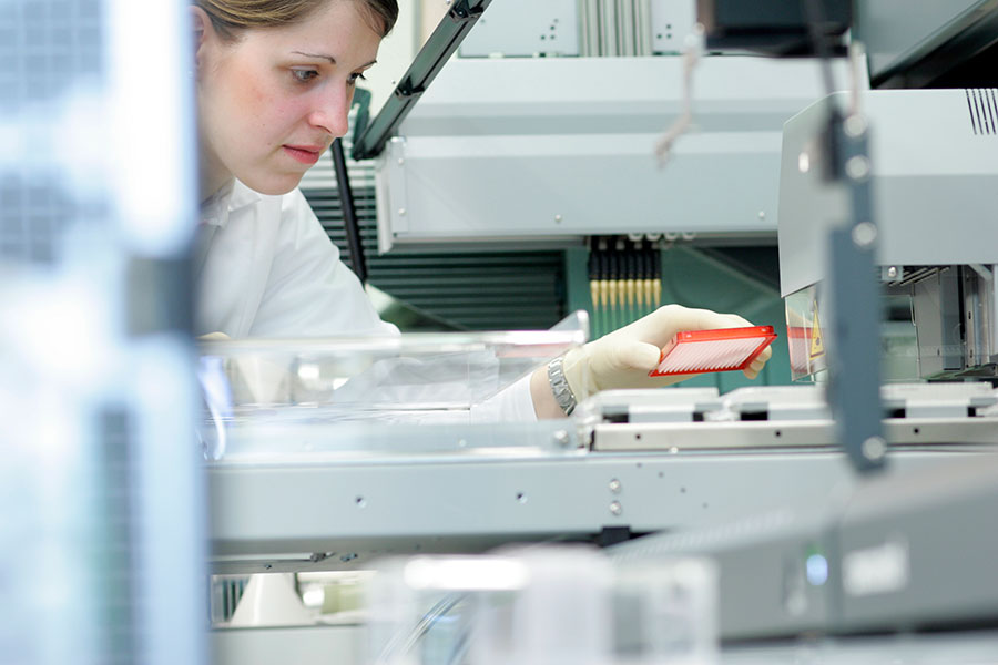 Eine junge Forscherin im Laborkittel blickt konzentriert auf ein großes graues Laborgerät, mit dem Proben analysiert werden können. Sie hält mit ihrer linken Hand eine rote rechteckige Platte mit kleinen Vertiefungen für Proben über die Mitte des Laborgerätes, um sie dort zu platzieren. 