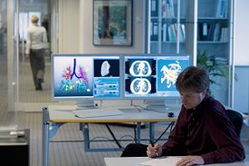 Ein Forscher macht sich Notizen, im Hintergrund sind zwei Monitore mit Ergebnissen der medizinischen Bildgebung