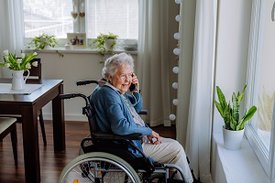 Eine ältere Frau sitzt in einem Rollstuhl vor einem Fenster und telefoniert mit einem Smartphone