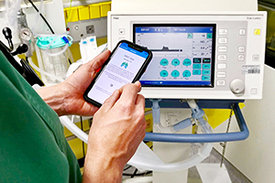 Ein Mitarbeiter auf der Intensivstation hält ein Mobiltelefon in den Händen und vergleicht die Daten einer medizinischen App mit einem Überwachungsgerät. 