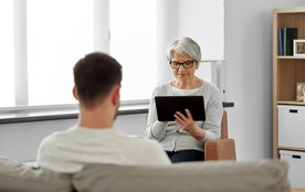Eine Frau blickt auf ein Tablet und spricht mit einem Mann, der ihr gegenüber sitzt.