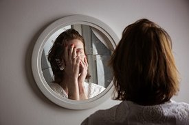 Eine Frau steht vor einem zerbrochenen Spiegel und hält sich die Hände vor das Gesicht