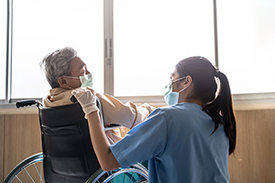 Älterer Patient im Rollstuhl mit asiatischer Pflegerin, beide tragen einen Mundschutz