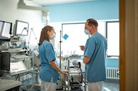Eine Frau und ein Mann in blauen Kitteln beraten sich in einem Krankenzimmer, das mit zahlreichen Geräten ausgestattet ist