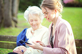 Junge Frau sitzt mit älterer Dame auf Parkbank und beide schauen auf ein Handy, dass die junge Frau in der Hand hält.