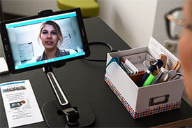 Das Bild zeigt ein Mobiltelefon, das auf einer Tischhalterung steckt. Auf dem Bildschirm des Telefons läuft eine Videokonferenz.