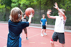 Kinder spielen mit Ball auf Sportplatz, wobei ein Junge ein Cochlea-Implantat hat und eine Hörgerät hinter dem rechten Ohr trägt.