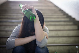 Mädchen sitzt auf einer verlassenen Treppe mit gesenktem Kopf in den verschränkten Armen und in einer Hand eine Flasche mit Alkohol haltend.