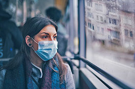 Dunkelhaarige Frau mit Mund-Nasen-Schutz sitzt in Zug und schaut aus dem Fenster