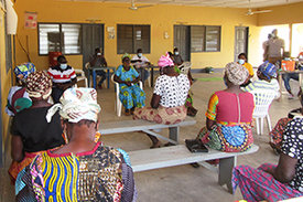Frauen aus Afrika sitzen mit dem Rücken zum Betrachter auf Holzbänken und hören einer Frau zu. 
